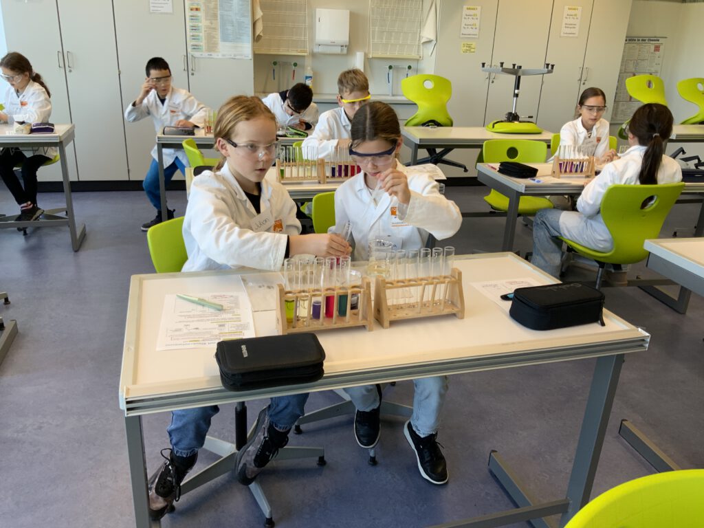 Schülerinnen und Schüler experimentieren mit Schutzbrillen und Laborkitteln im naturwissenschaftlichen Unterricht